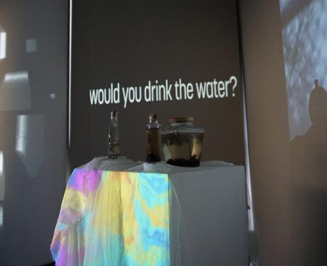 Photo of the art exhibit. A display with jars of natural water, with colorfully lit cloth under it. Background wall reads, "Would you drink the water?" in reference to the water contaminated in the Ohio train derailment.
